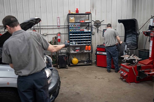Holiday Road Trip Safety & Fun | Lonsdale Auto Works Tips. Two male mechanics at Lonsdale Auto Works checking fluids on vehicle and balancing tire in shop.