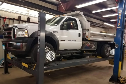 Wheel alignment services near me at Lonsdale Auto Works in Lonsdale, MN. Image A white Ford F550 truck is raised on a hydraulic lift inside a well-lit garage undergoing wheel alignment